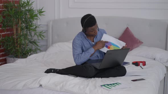 Wide Shot Portrait of Confident African American Young Man in Earphones Sitting on Bed Pointing at