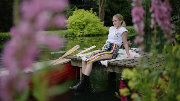 A Beautiful Woman Sitting On The Wooden Jetty By The Lake With A Boat Floating - full shot