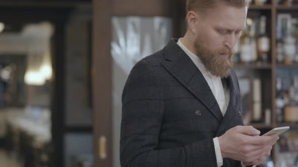 Portrait of a Successful Young Bearded Confident Businessman Typing on a Cell Phone While Standing
