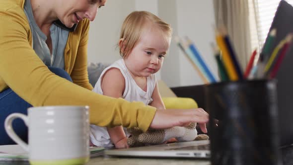 Caucasian mother holding her baby using laptop and talking on smartphone while working from home