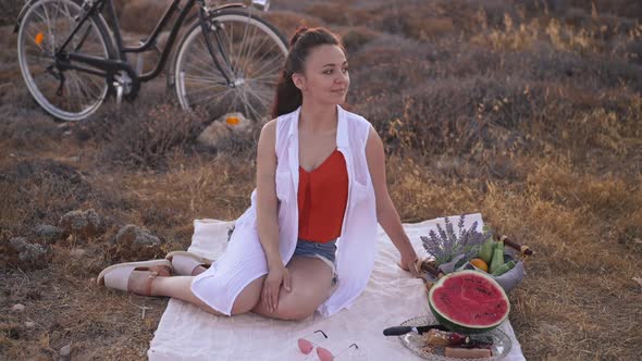 Wide Shot Relaxed Smiling Young Woman Sitting on Blanket with Fruits Looking Around