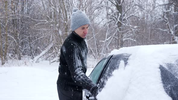 Man in a Black Sweater and a Gray Hat Cleans the Car From Snow During a Snowfall in Slow Motion