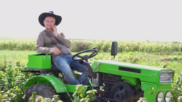 Young Farmer Sitting on a Tractor and Talking on a Smartphone in the Field