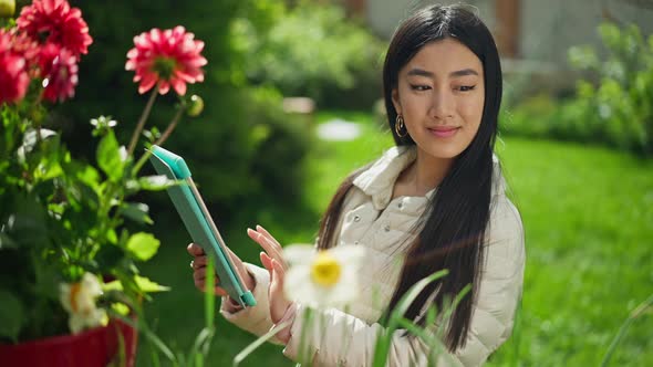 Confident Smiling Asian Woman Typing on Tablet Looking at Blooming Flowers in Sunny Summer Spring