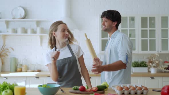 Young Couple Laughing Holding Kitchenware Singing Song Together Dancing To Music Enjoying Cooking in