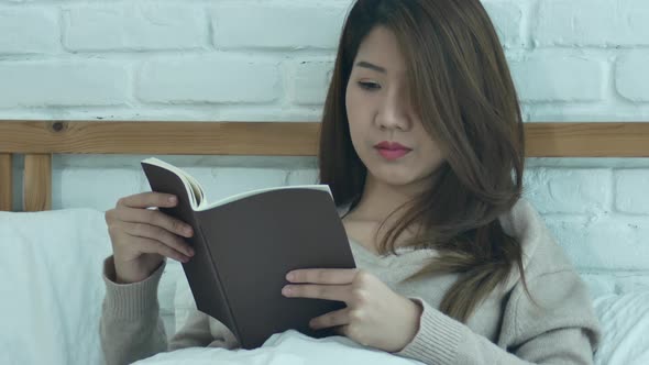 sian woman enjoying lying on the bed reading book pleasure in casual clothing at home.