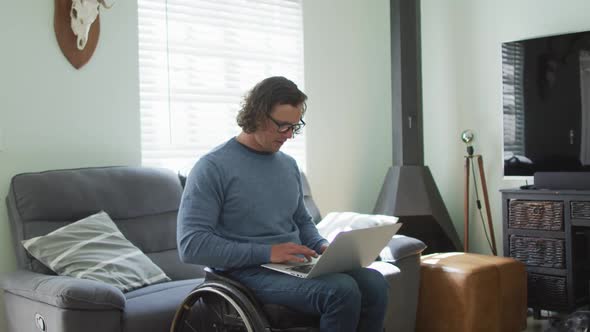 Happy caucasian disabled man in wheelchair using laptop in living room