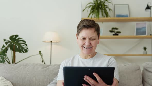 Smiling Elderly Mature Woman Resting on Sofa Using Digital Tablet Alone at Home