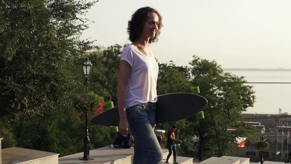 Young Attractive Girl Walking Holding Her Longboard in the City Street During Sunrise Slow Motion