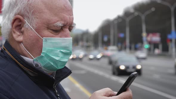 Elderly Man In Virus Proof Mask Uses Phone While Standing By Road With Traffic