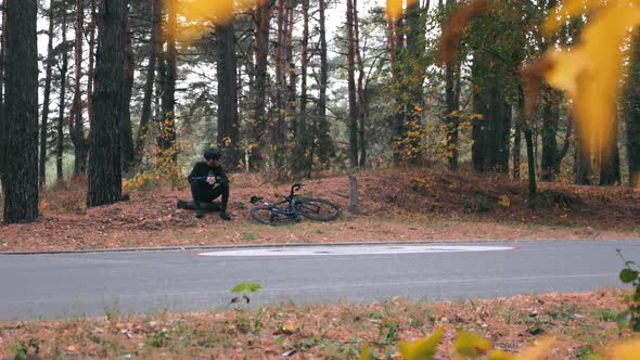 Male cyclist sitting on log in autumn park and drinks water after intensive cycling training