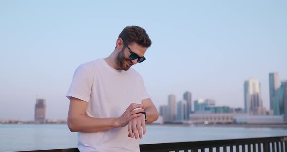 A Young Man Standing on the Waterfront in the Summer Uses the Smart Watch Screen