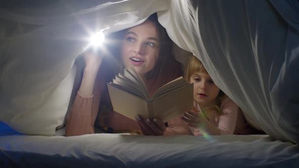 Mother and Daughter Lying in Bed at Home Under Duvet Blanket and Reading Bedtime Stories Fairytales