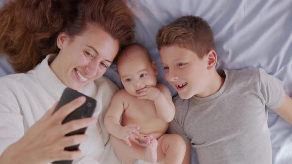 Happy Mother with Her Kids Are Making a Selfie or Video Call To Father or Relatives in a Bed