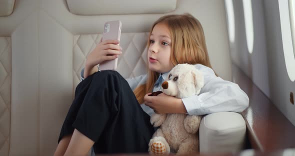Adorable Little Girl with Teddy Bear Talking on Video Chat on Smartphone Traveling By Airplane