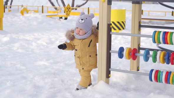 Toddler in yellow warm clothes 12-17 months to learn to walk in the snow on the playground.