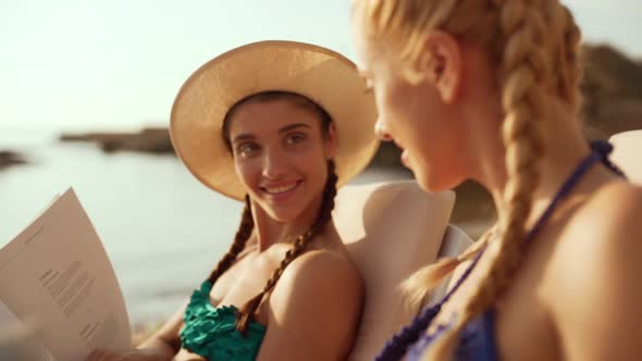 Two Beautiful Girls at the Seaside One Reading Magazine Another with Book Talking Discussing