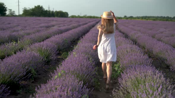 Blonde Girl Running Along the Lavender Field in a White Dress and a Straw Hat in Summer