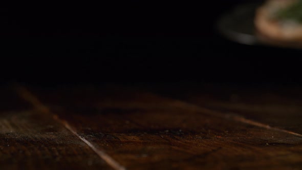 Camera follows putting a white pizza with prosciutto and arugula on a table. Slow Motion.