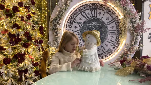 A Child Girl Play with a Doll Snowman in Near a Christmas Tree with Big Clock