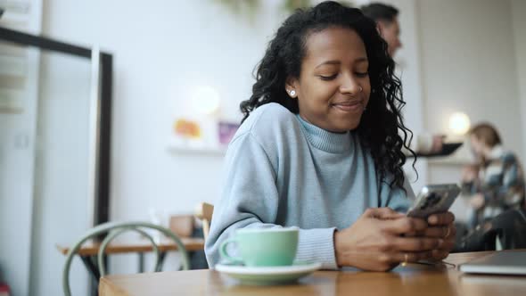 Happy African woman wearing blue sweater texting on phone and looking to the side
