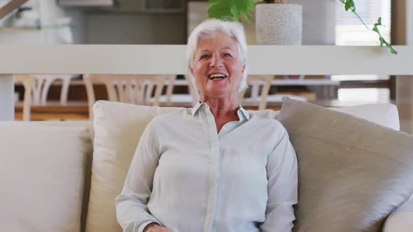 Portrait of senior caucasian woman talking and waving looking at camera while sitting on couch at ho