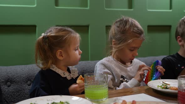 Kid Girl Play in Colorful Antistress Toy Popit and Little Toddler Girl Eating Pizza at Cafe