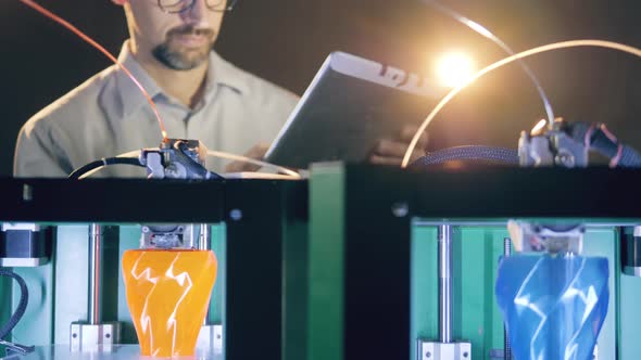 Two 3D Printers Work with Vases While a Man Controls Them. Engineer Working with 3D Printer