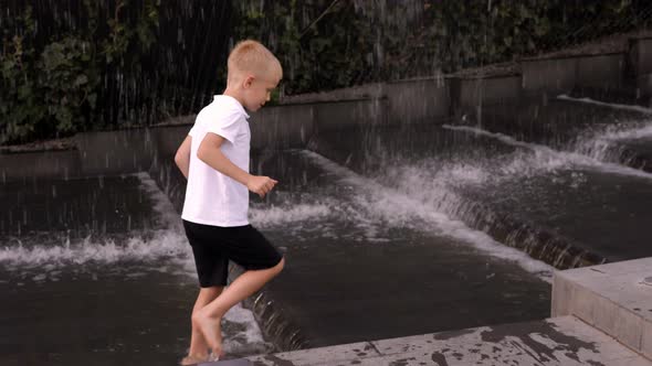 A Little Boy Plays in a Ground Fountain with a Waterfall in a City Park