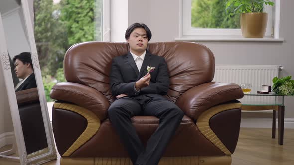 Wide Shot Portrait of Serious Thoughtful Young Asian Man in Wedding Suit with Boutonniere Sitting on