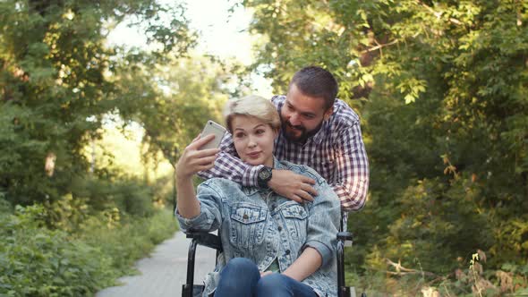 Disabled Person Caucasian Woman in Wheelchair with Her Beloved Man in Park Takes Selfie on Phone for