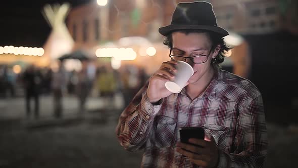 Young Man Drinking Coffee And Using Phone At Street In Evening