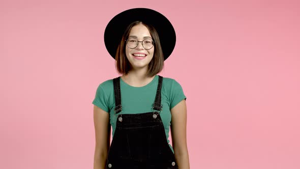 Young Smiling Woman in Hat, Glasses and Overall Looking To Camera. Portrait of Cute Beautiful Girl