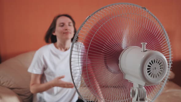 Young Woman Sits in Front of Electric Fan Waving Hands Air Stream Blowing Hair