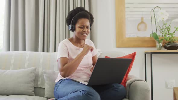 Happy african american woman wearing headphones sitting on sofa, using laptop