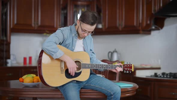 Young Romantic Caucasian Man in Eyeglasses Playing Guitar Sitting on Kitchen Table at Home