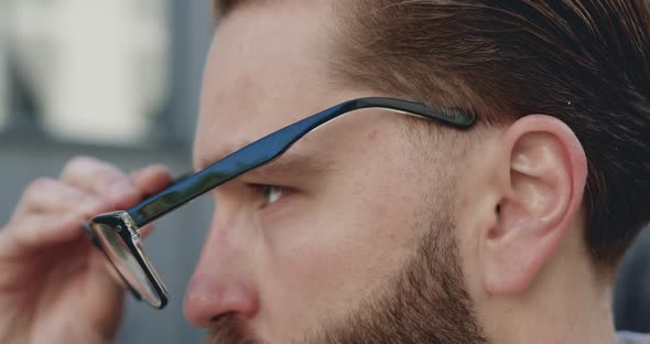 Young Bearded Man which Putting on His Glasses Poses on Camera Outdoors