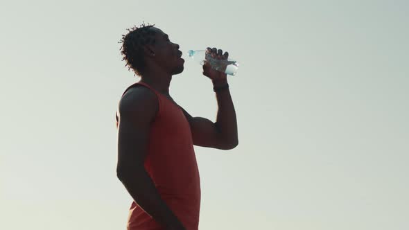 African-American man drinks water from sports bottle after jogging outdoors, side view. Athlete rest