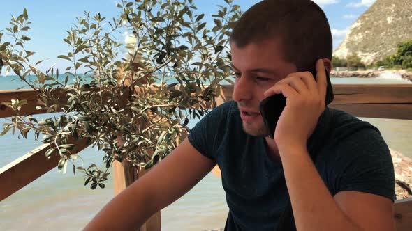 Slow zoom of a young handsome man with beard leading a conversation through the phone with the sea i