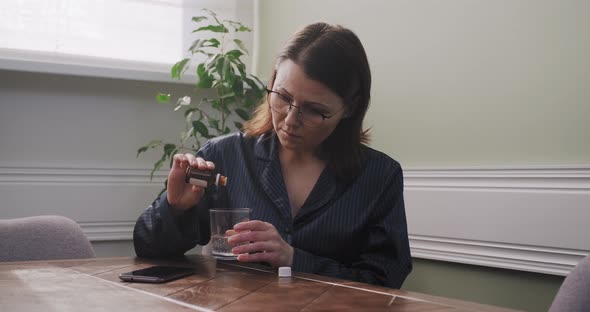 Mature Woman Dripping Drops with Glass of Water, Drinking Sedative Medicine, at Home in Pajamas