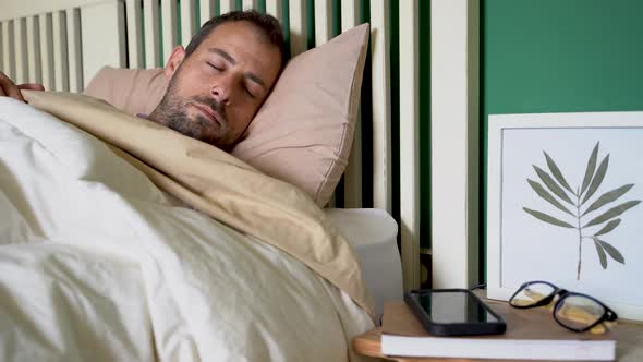 Man waking up in bed, switching off alarm on smartphone