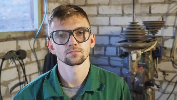 Exhausted Young Repairman in Protective Glasses Looking Into Camera at Garage