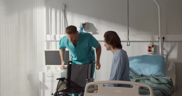 Man Nurse Helping Disabled Young Patient Sit in Wheelchair in Hospital Ward