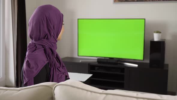 A Young Beautiful Muslim Woman Watches TV with Green Screen and Nods at the Camera with a Smile