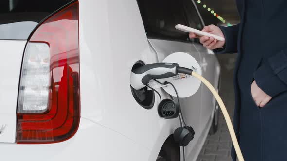 Woman Unplugging the Power Supply Cable From an Electric Car and Using Her Smartphone for Unlocking