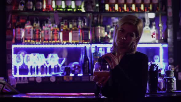 Beautiful Girl with Blond Hair Standing Near the Bar Counter on the Background of Flickering Light