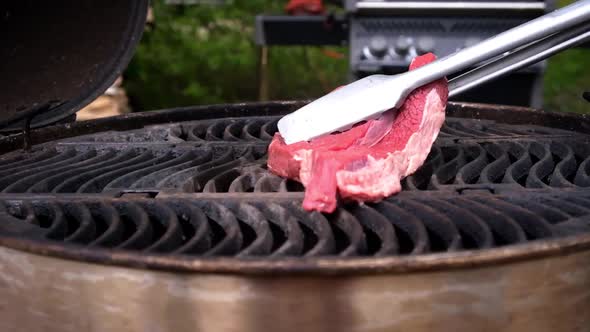 A piece of fresh red meat is put on a hot grill with tongs
