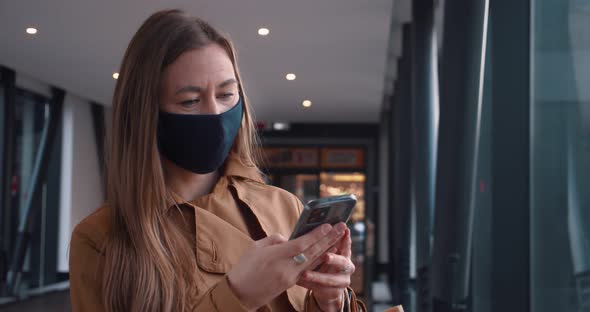 Beautiful Young Serene Shopper Woman in Face Mask Using Smart Phone News Reading App at Mall Passage