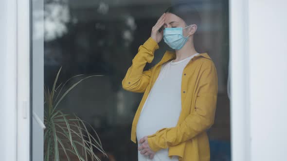 Stressed Worried Pregnant Woman in Covid19 Face Mask with Head Ache Standing Indoors Looking Out