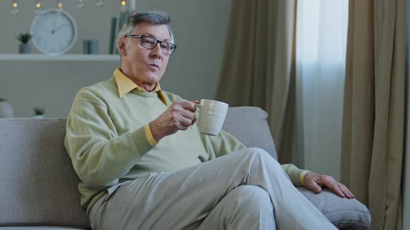 Old Man Wearing Glasses Sit on Couch in Living Room Hold Hot Drink Cup Pensive Elderly Grandpa